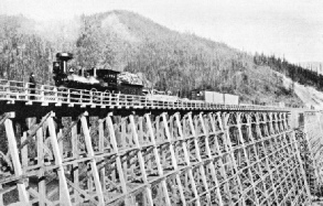 EARLY WOOD-BURNING LOCOMOTIVE hauling trucks across a wooden trestle bridge in the Selkirk Mountains, Canadian Pacific Railway